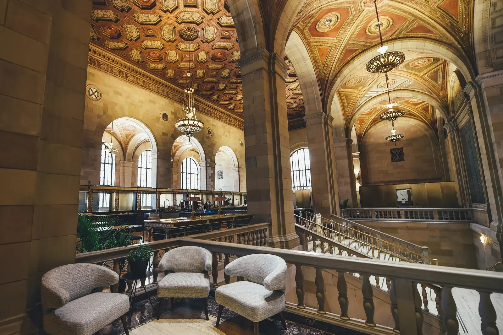 A grand banking hall with a gilded coffered ceiling, arched windows, brass chandeliers and armchairs on the gallery