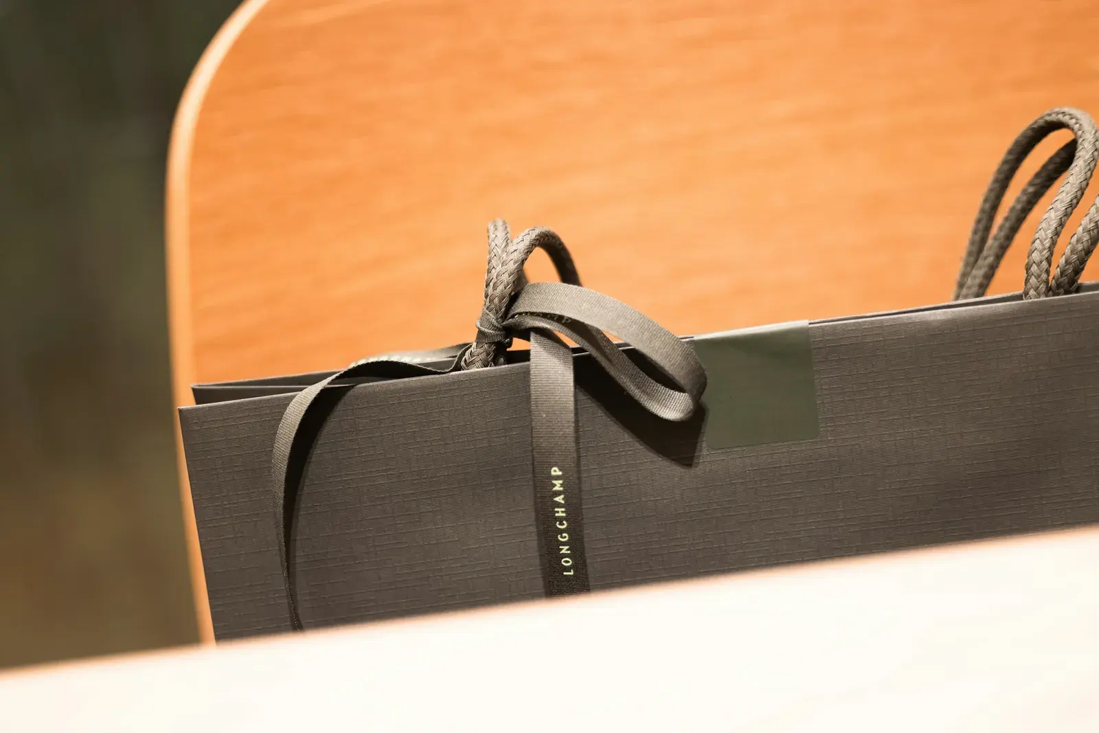 A dark grey Longchamp shopping bag tied with a branded ribbon, resting against a wooden chair
