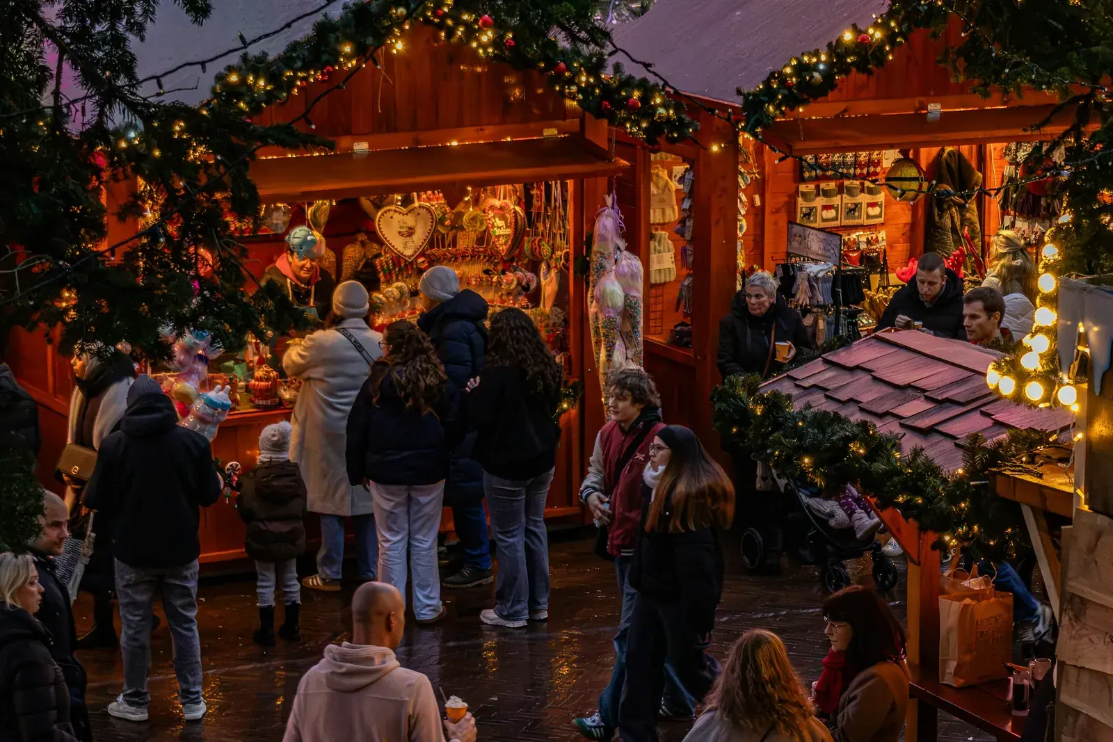 Shoppers browsing wooden Christmas market stalls hung with garlands, fairy lights and gingerbread hearts at dusk
