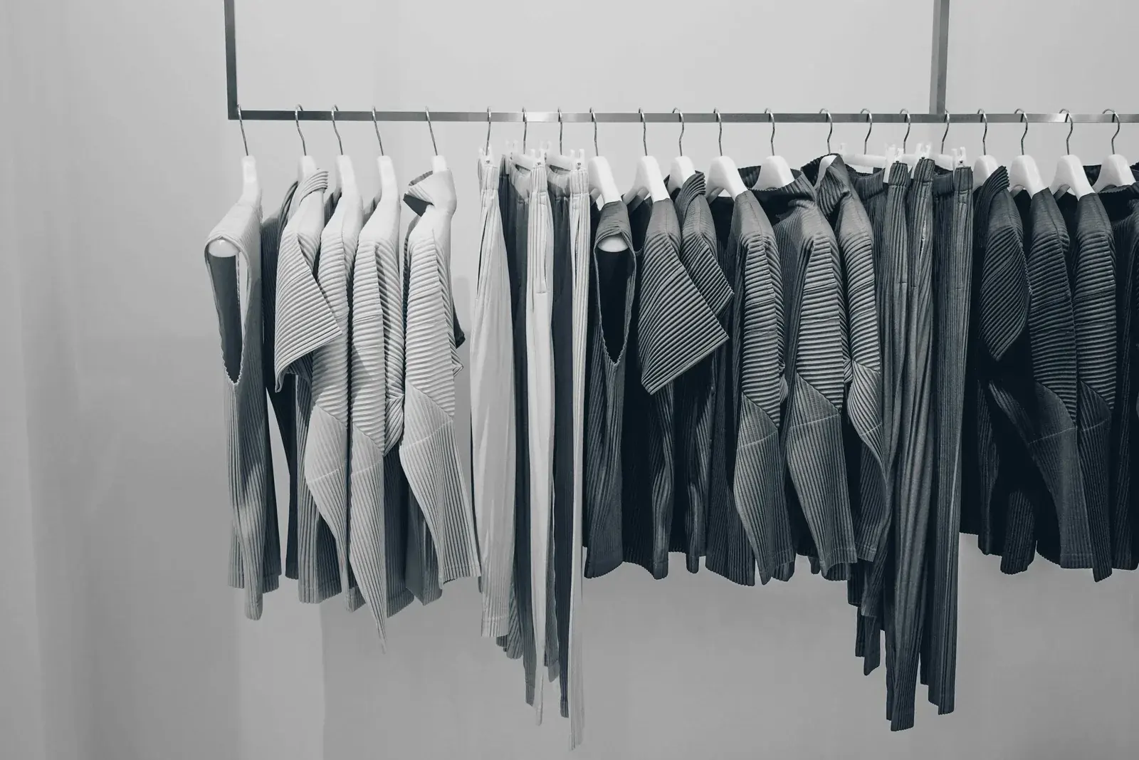 A short rail of pleated tops and trousers in white, grey and charcoal against a pale wall