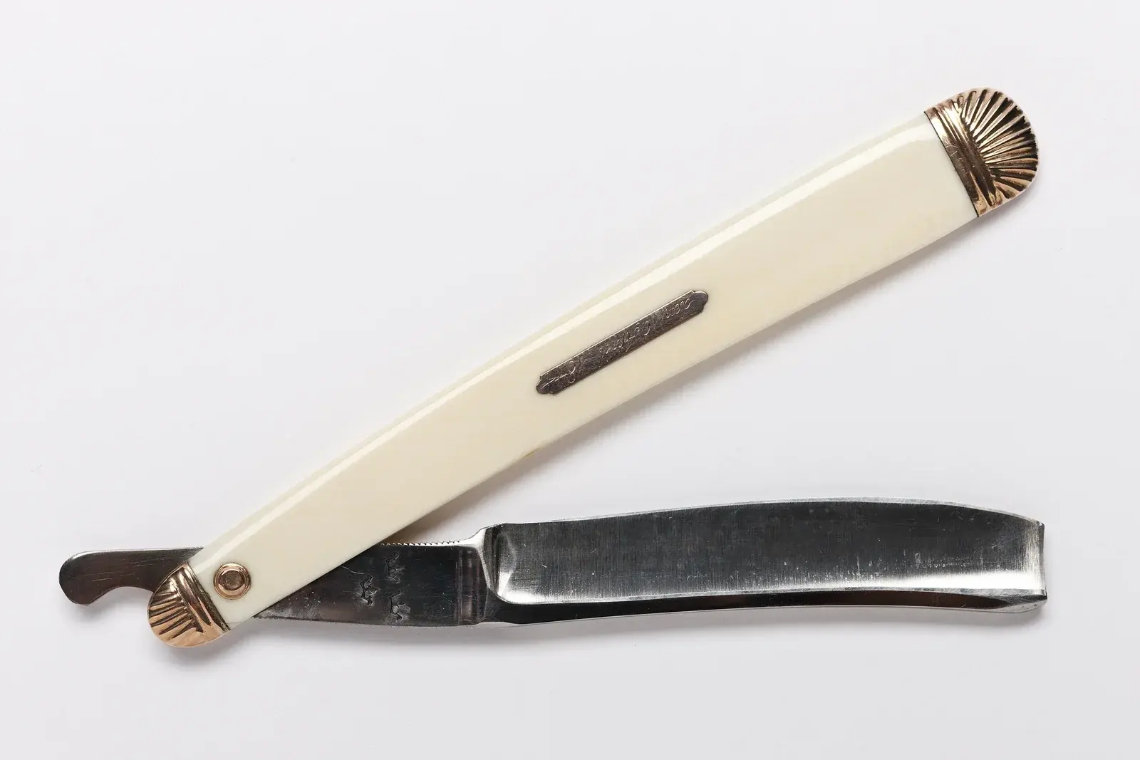 An antique straight razor with a cream handle and fluted gold-tone end caps, opened on a white surface