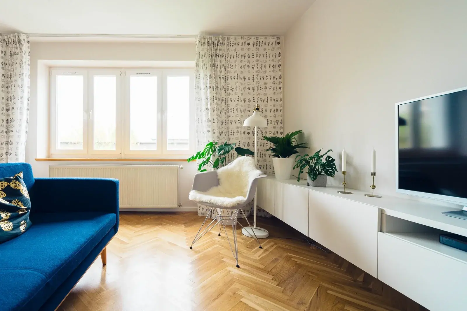 A bright Scandinavian living room with a blue sofa, herringbone oak floor, sheepskin chair, plants and a low white sideboard
