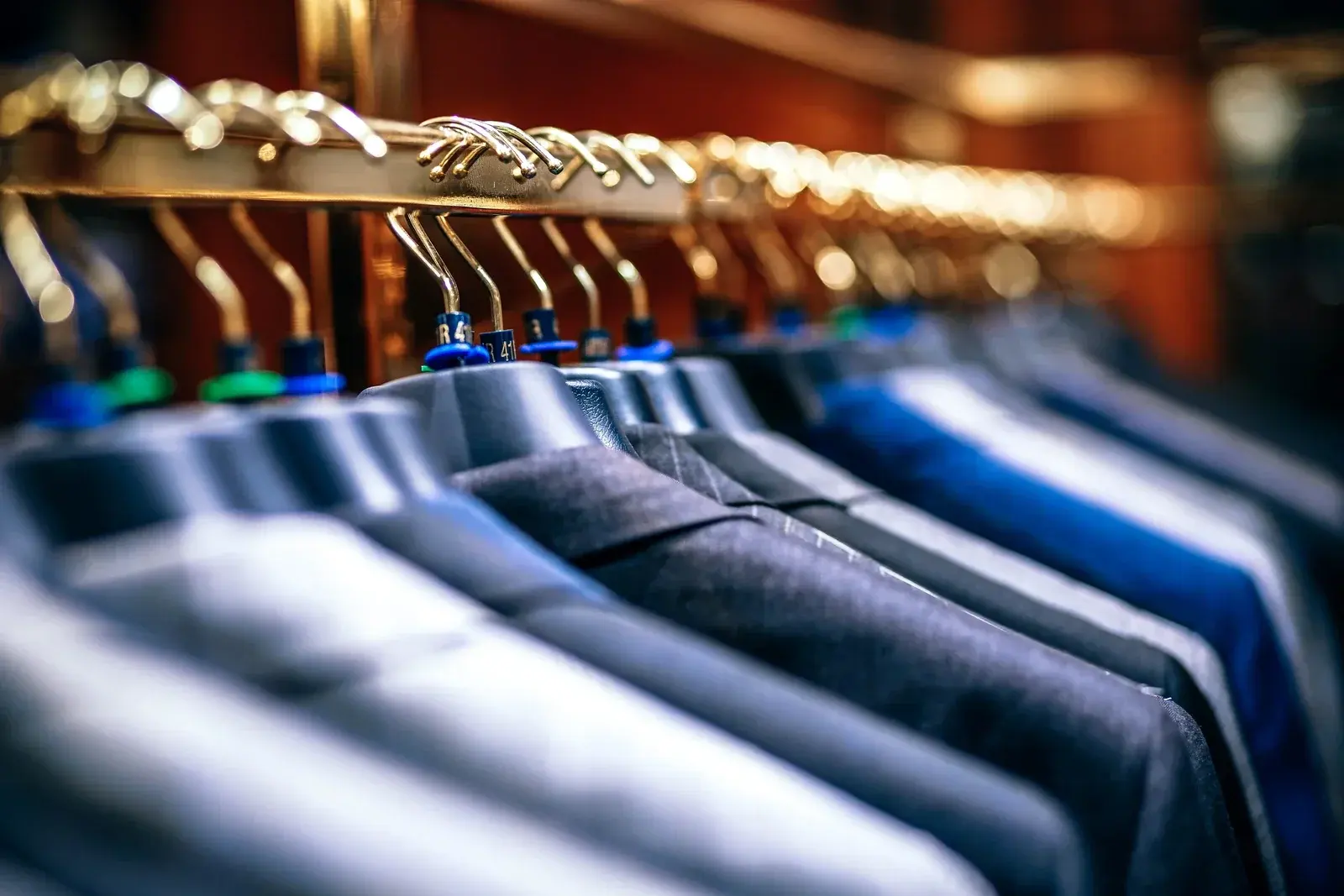 Grey and navy suit jackets hanging from a brass rail in a softly lit tailor's shop
