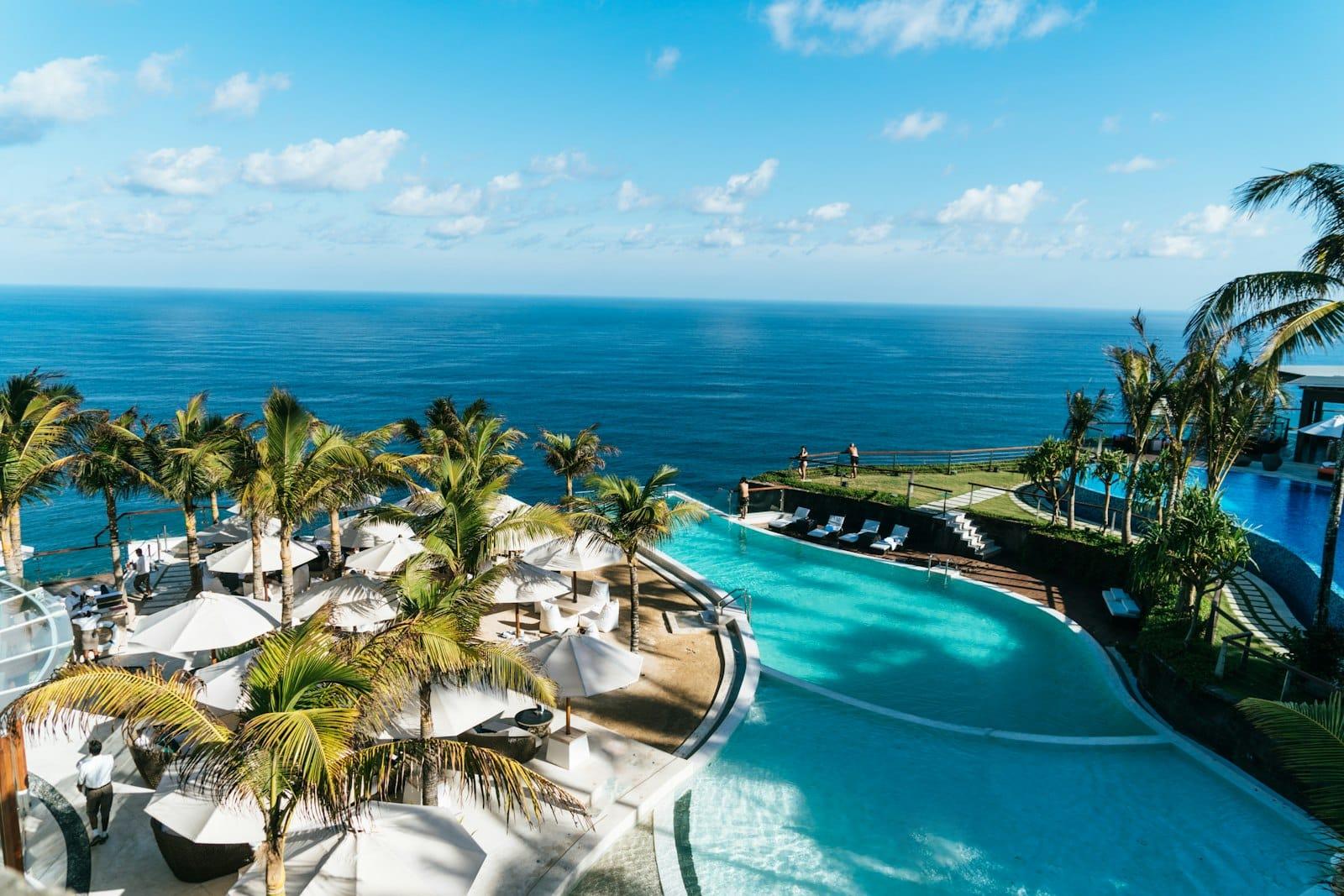 A cliff-top resort at midday: palms, white parasols and a turquoise pool above open sea