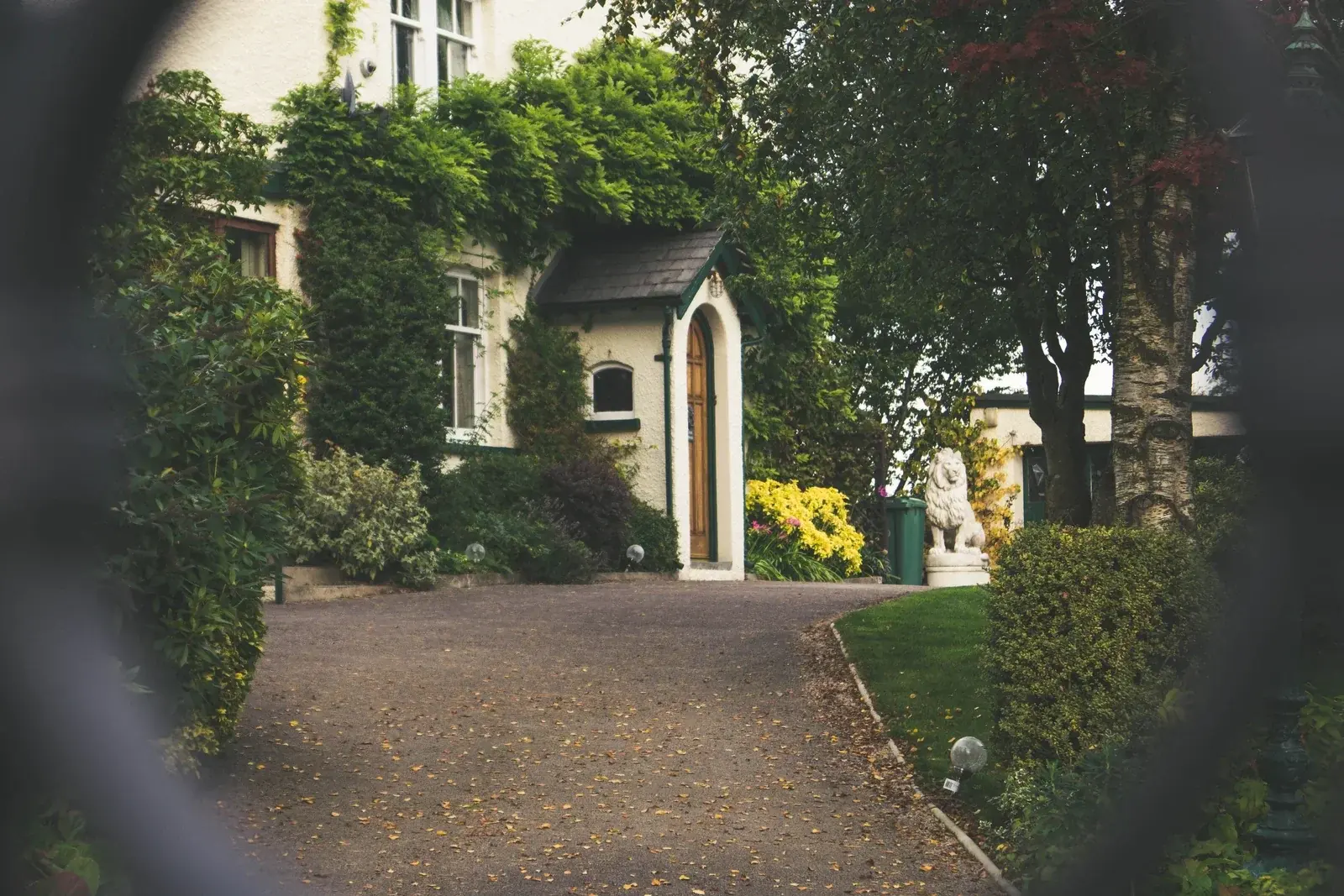 An ivy-covered country house with an arched front door and a stone lion, seen through the bars of a gate