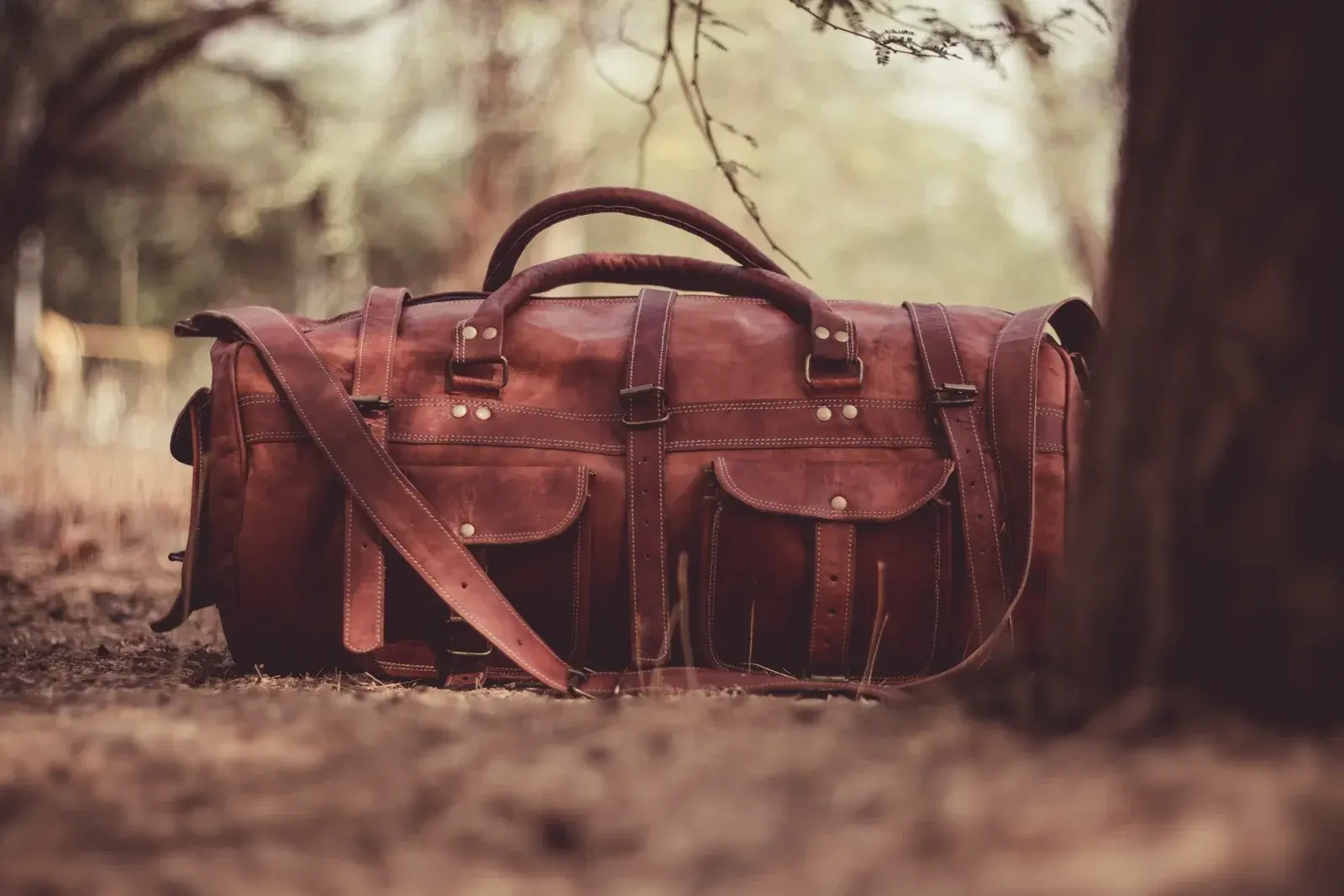 A well-worn brown leather holdall with buckled straps resting on a woodland path