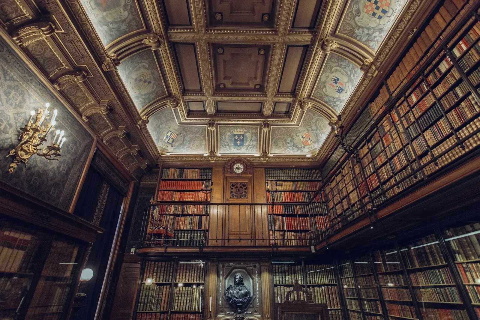 A wood-panelled private library with a coffered heraldic ceiling, a gallery of leather-bound books and a brass chandelier
