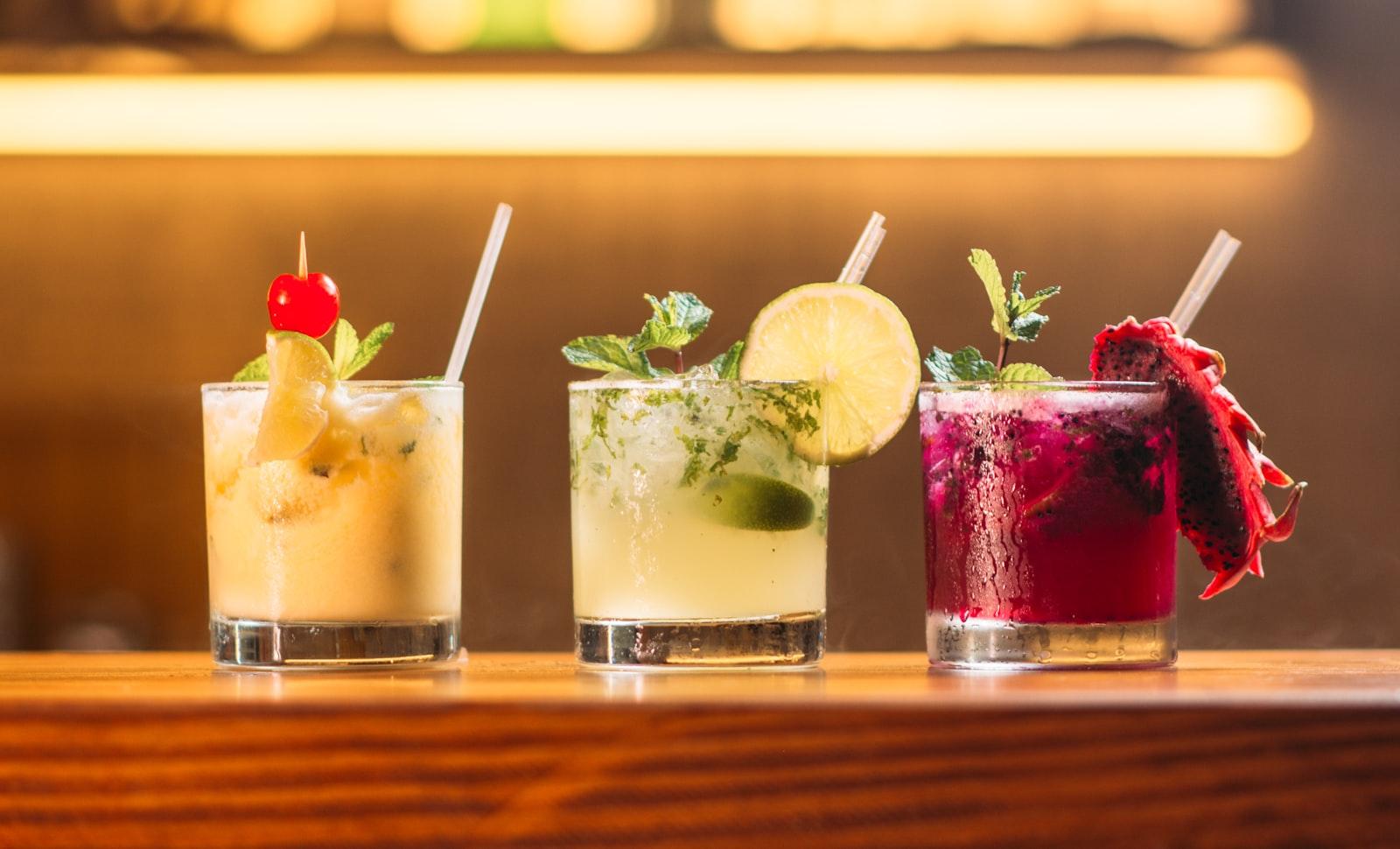 Three garnished cocktails in tumblers lined up on a lit wooden bar