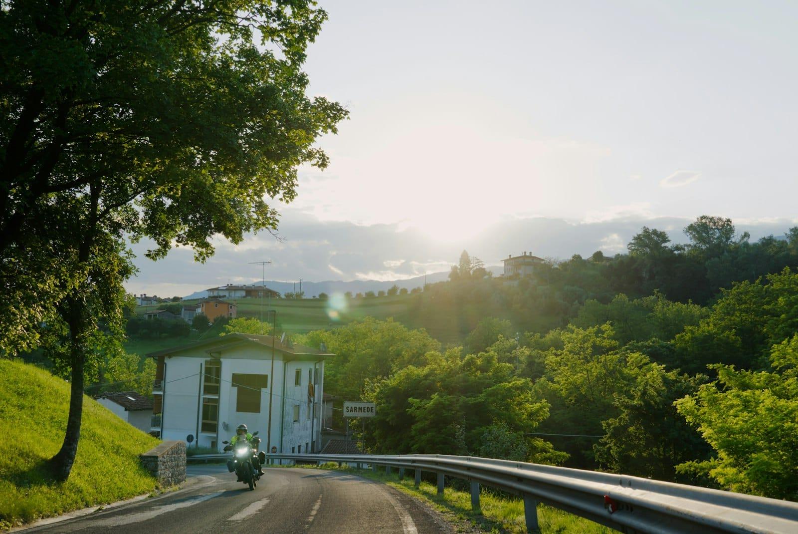 A motorcyclist rides into a hillside village at sunset, green slopes and houses beyond