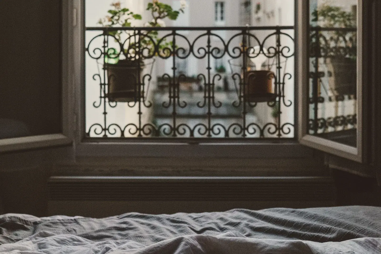 An unmade bed with grey linen before tall open French windows and a wrought-iron balcony in Paris