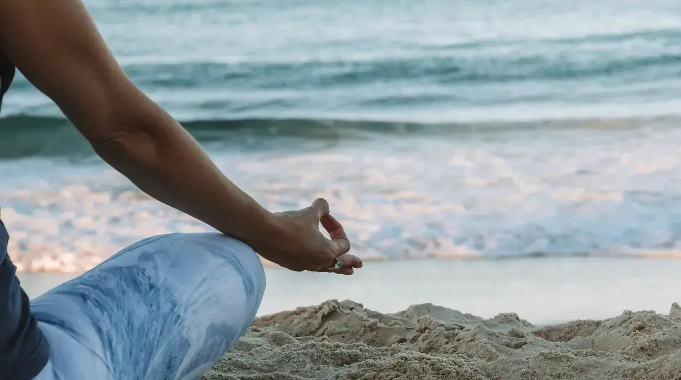 A hand resting on a knee in a meditation seat on the sand, surf breaking behind