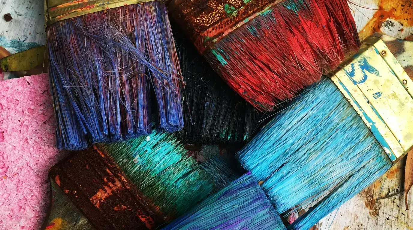 A pile of paint-loaded brushes in blue, red and turquoise beside a pink sponge.