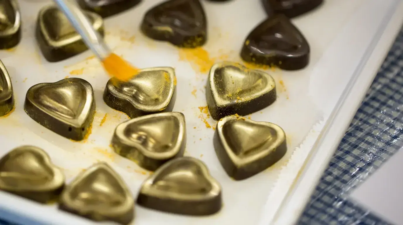 Heart-shaped chocolates being brushed with gold dust on a lined tray.