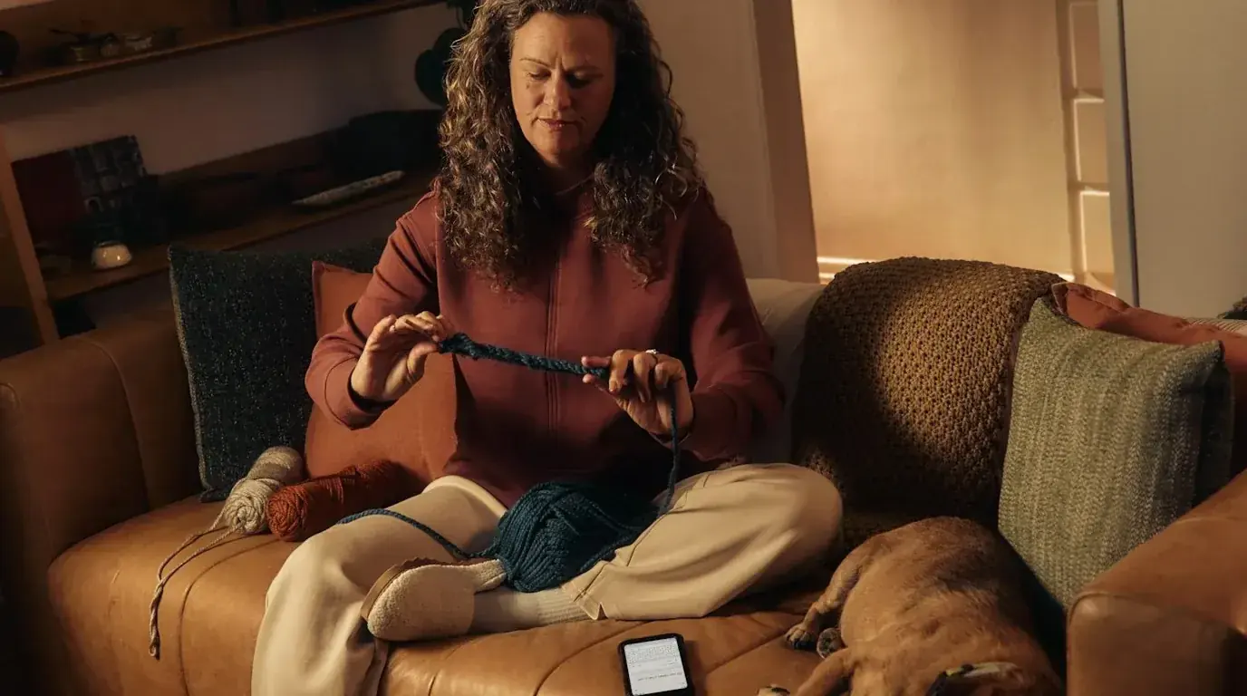 A woman knitting with dark teal yarn on a sofa in warm lamplight, a dog asleep beside her.