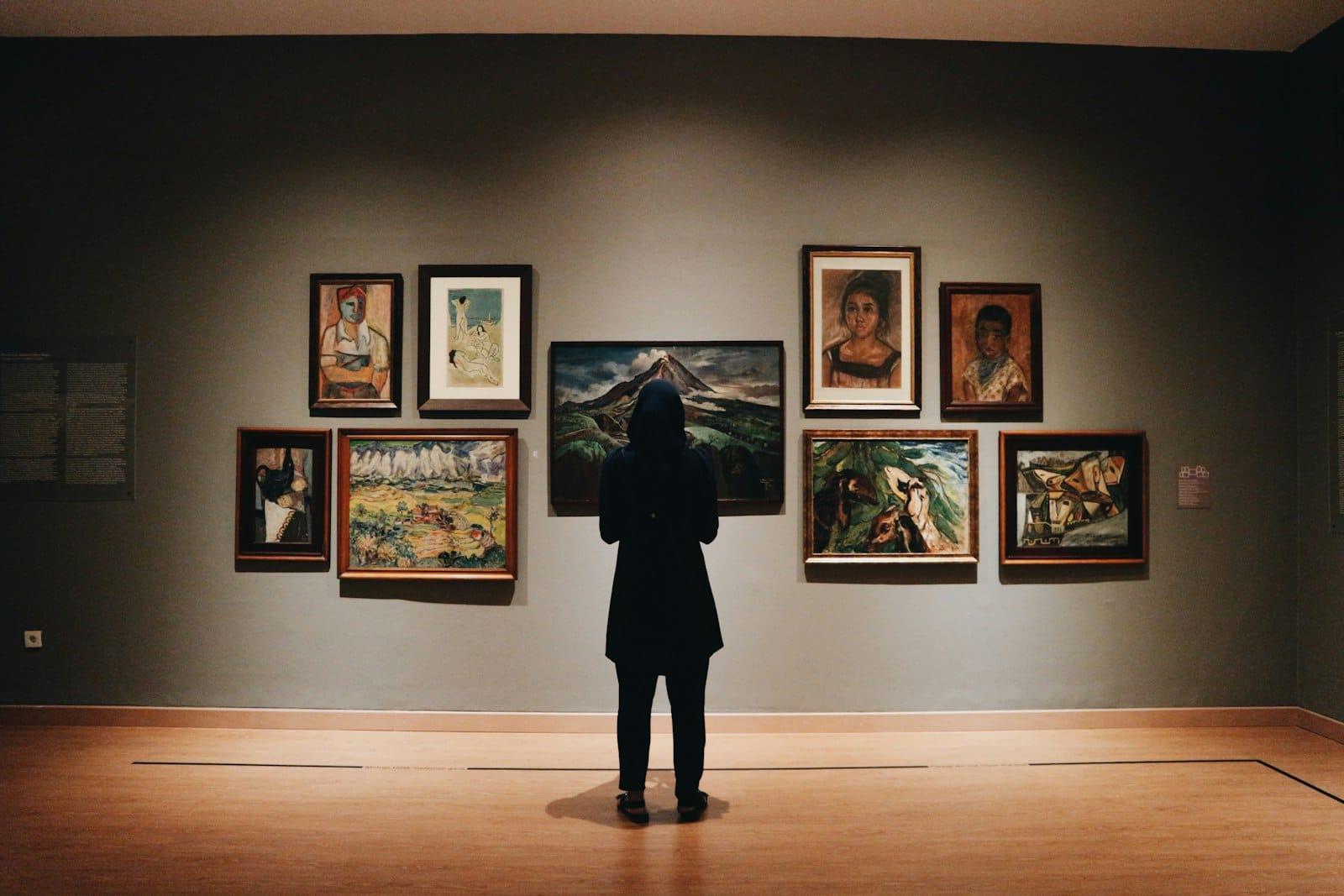 A lone visitor stands before a wall of framed twentieth-century paintings in a dim gallery