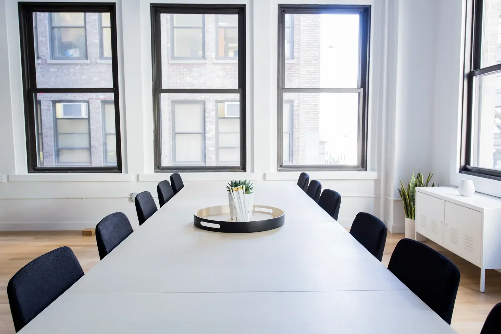 A long white boardroom table with black chairs beneath tall loft windows