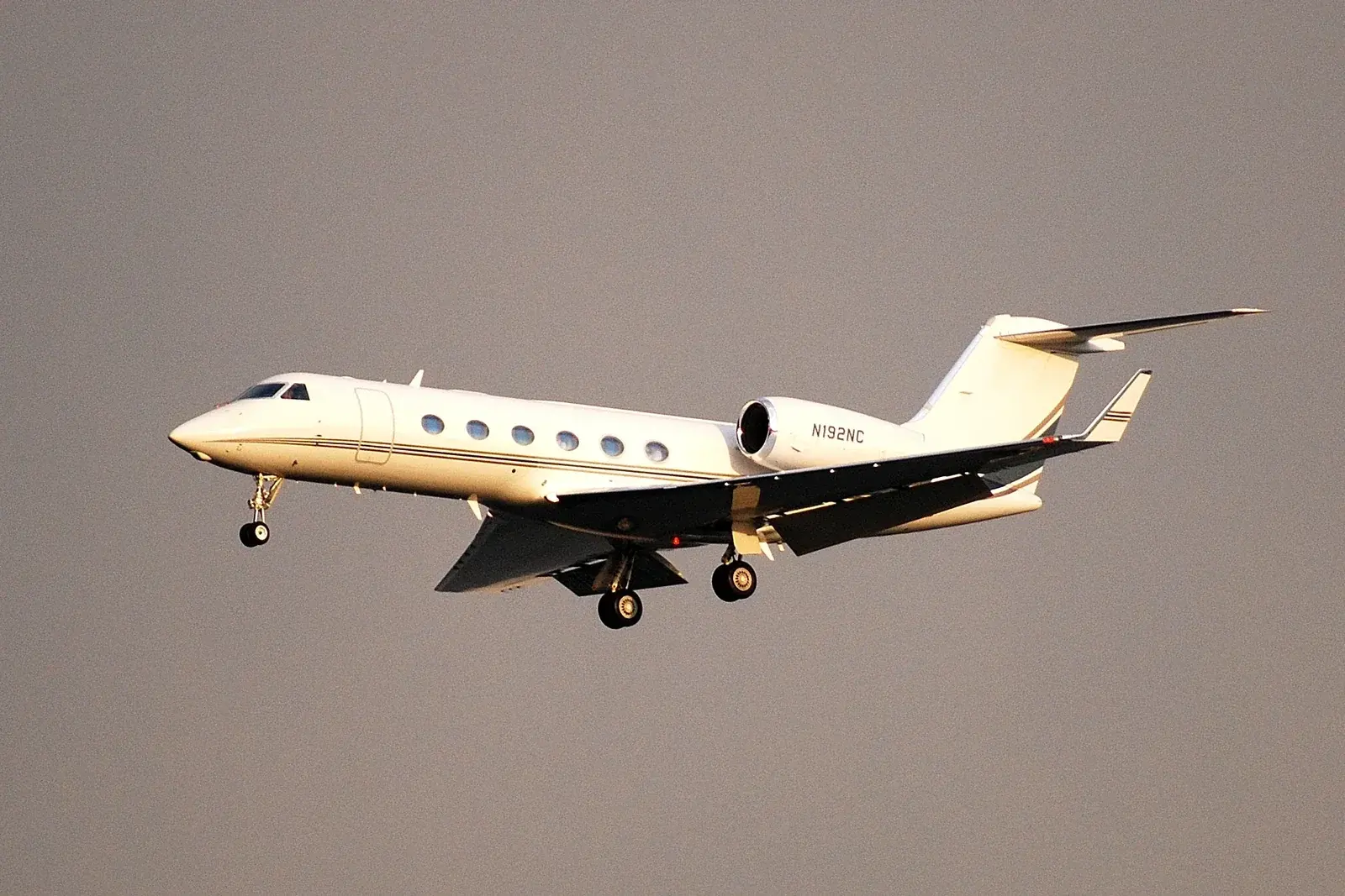 A white long-range business jet on final approach with its landing gear down, lit by low evening sun