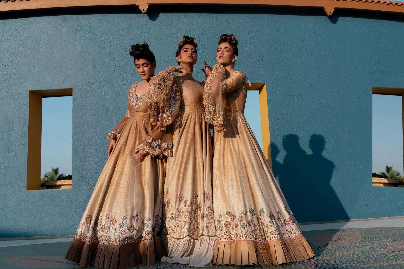 Three models in embroidered champagne silk gowns posed against a blue wall