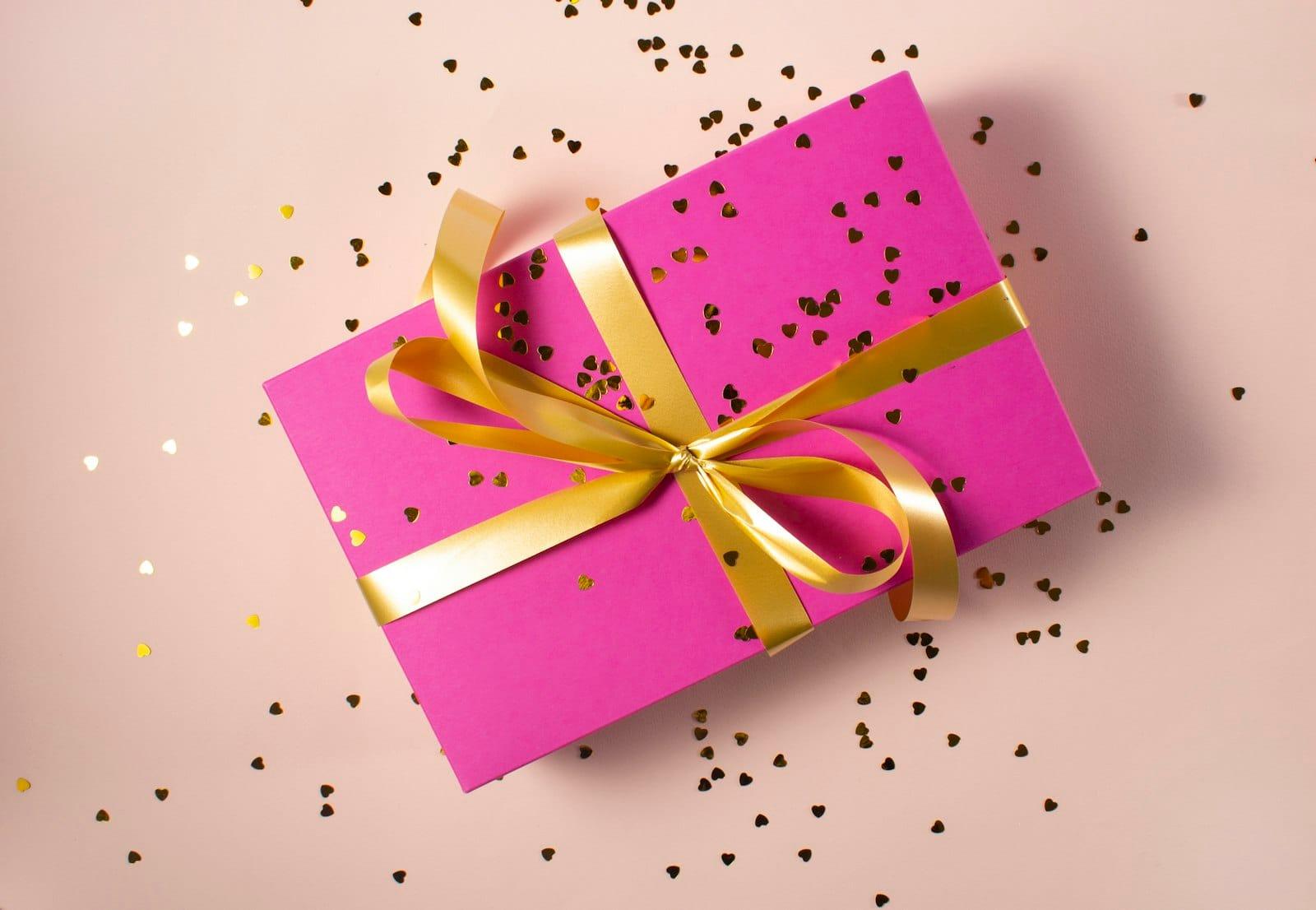 A magenta gift box with a gold ribbon on pale pink, gold heart confetti scattered around it