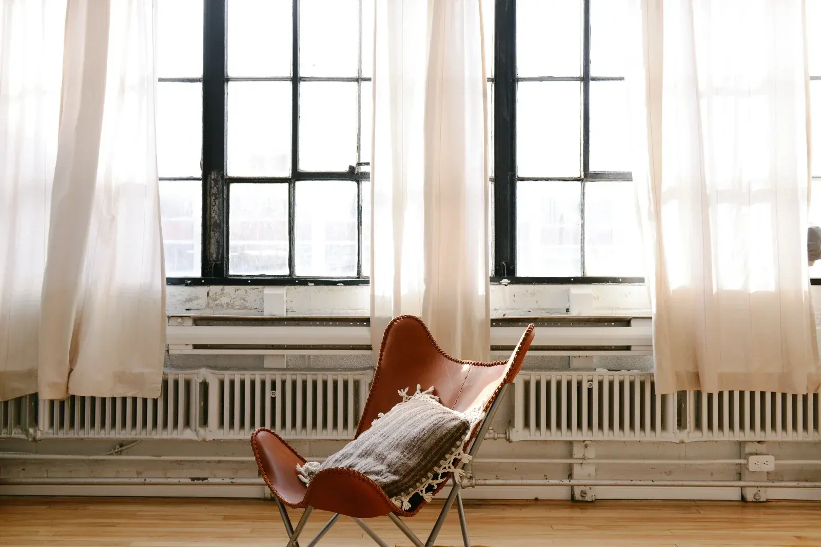 A tan leather butterfly chair with a fringed cushion before tall loft windows hung with sheer linen curtains