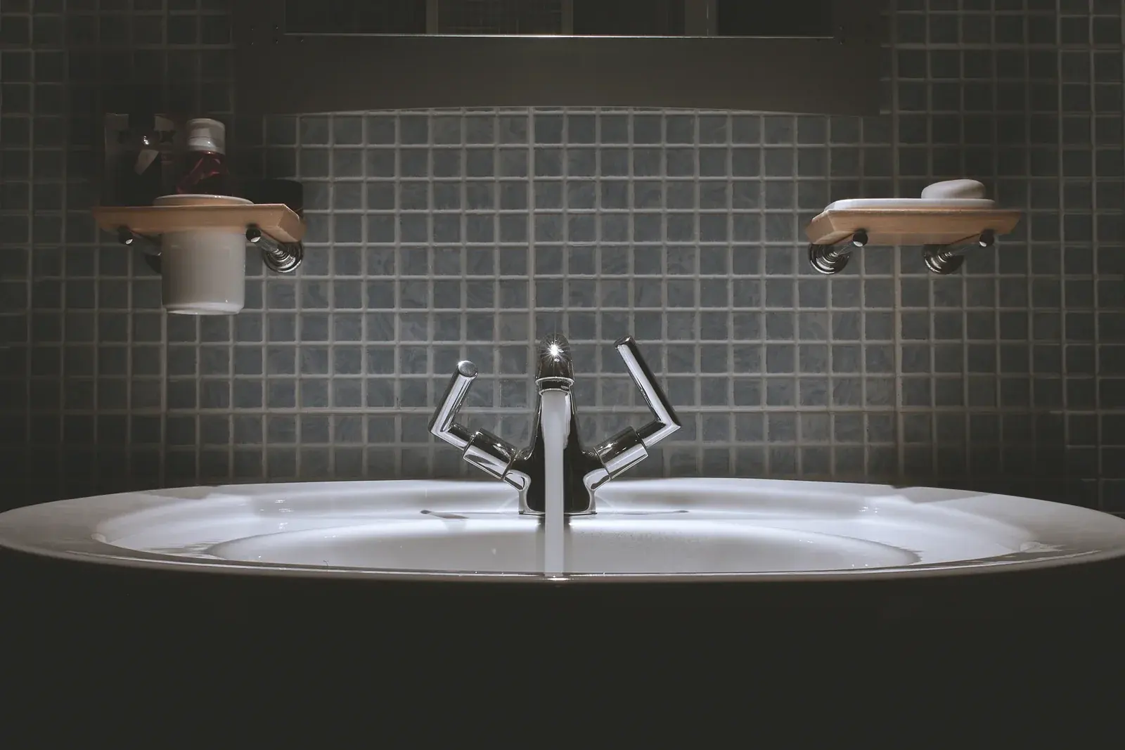 A chrome mixer tap running into a white basin against dark blue mosaic tiles, a soap dish on a wooden shelf