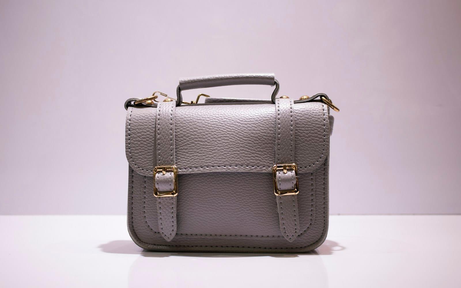 A grey leather satchel with gold buckles on a white surface against a lilac wall