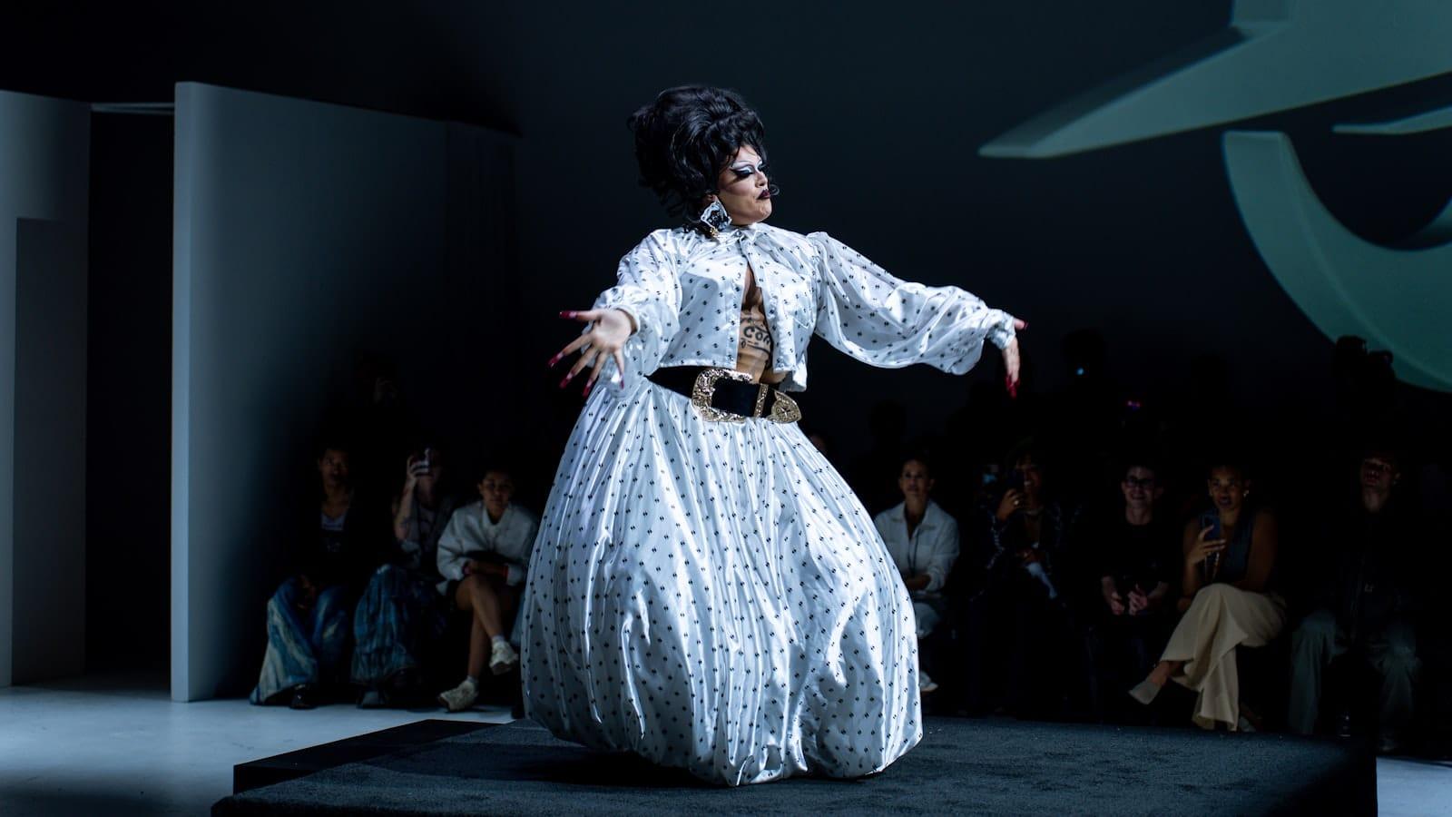 A performer on a runway in a voluminous white polka-dot gown, the audience seated in the dark