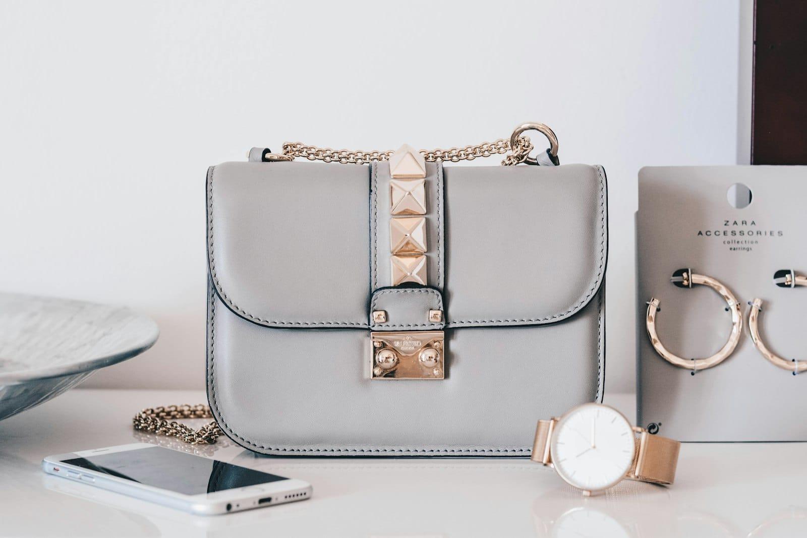 A grey studded leather shoulder bag with a gold chain, a watch and a phone beside it