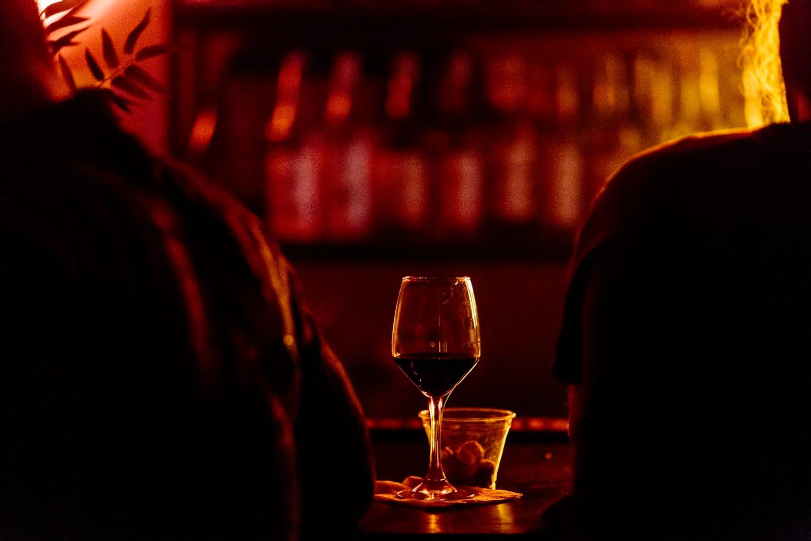 A glass of red wine on a bar counter between two silhouetted drinkers, bottles behind